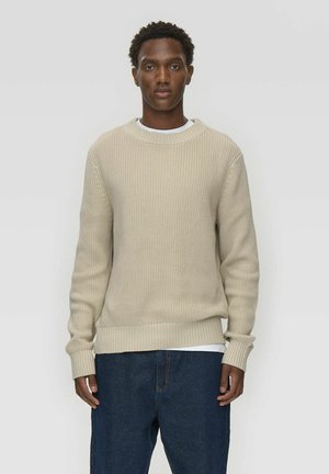 Beige knitted sweater with a round neck, ribbed cuffs, and hem, layered over a white shirt. Features textured pattern on the body.