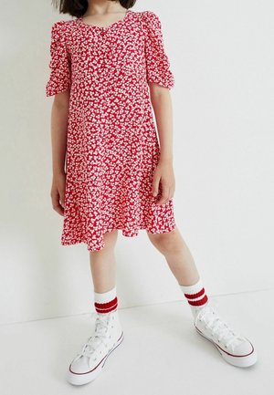 Czerwona sukienka w biały kwiatowy wzór, z bufkami i rozkloszowanym dołem. Połączona z białymi sneakersami na wysokiej podeszwie i skarpetkami w czerwone paski.