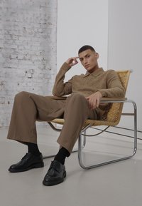 Polo shirt a maglia marrone testurizzata, pantaloni beige a gamba larga e scarpe di pelle nere. Seduto su una sedia intrecciata con un telaio in cromo, contro un muro bianco.
