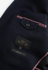 Granatowa marynarka o fakturowanej tkaninie, w slim fit, z kontrastowym podszewkowym kieszonką w kwiatowy wzór. Metka pokazuje markę i rozmiar.