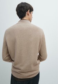 Jasnobrązowy dziany sweter o subtelnej fakturze i kołnierzu, pokazany od tyłu, zestawiony z ciemnymi dżinsami.