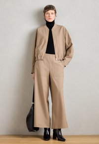 Beige bomberjacka med elastisk nederkant, svart polotröja, vida beiga byxor och svarta klackstövlar. Håller en svart väska med strukturerat mönster.