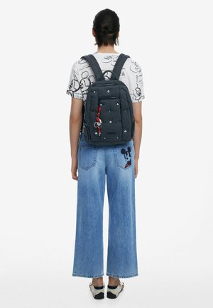 Grijze stoffen rugzak met gevoerde banden, voorvak met ritssluiting en decoratieve sleutelhanger. Model draagt een grafisch T-shirt en wijde denim jeans.