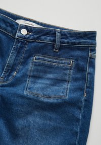 Denim jeans in a medium blue wash, featuring a five-pocket design, copper stitching, and a front button closure with a small logo tag.