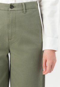 Ingrandimento della persona che indossa pantaloni verdi a vita alta con bottone e una camicia bianca a maniche lunghe, mano destra rilassata al fianco.