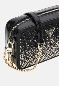 Črna sintetična torbica z učinkovito obliko, ki ima bleščeč gradient iz rhinestones in zlato verižico. Zadrga na vrhu.