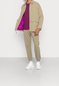 Beige zip-up jacket with purple lining, paired with a purple t-shirt and beige joggers. White sneakers complete the outfit.