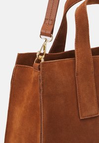 Brun ruskinds tote taske med en tekstureret overflade, der har en trykknaplukning og en aftagelig læderrem, dekoreret med en guldfarvet krog.