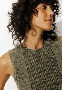 Femme aux cheveux noirs bouclés portant un haut sans manches en tricot vert olive avec des motifs texturés, de profil, devant un fond neutre.