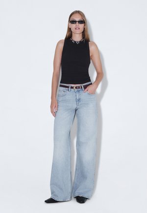 Vrouw met een zwart mouwloos topje, lichtblauwe wijde jeans, zwarte riem, zwarte puntige schoenen, zonnebril en een ketting, staand tegen een witte achtergrond.