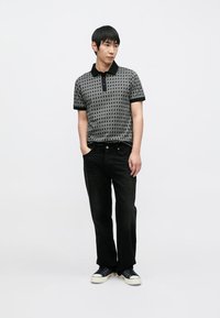 Polo em preto e cinza com um padrão geométrico, apresentando uma gola preta e uma carcela com dois botões, combinado com jeans preto e sapatilhas.