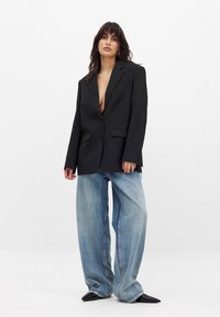 Schwarze oversized Blazerjacke mit Revers und zwei vorderen Taschen, kombiniert mit weit geschnittenen hellblauen Jeans und spitzen schwarzen Schuhen.