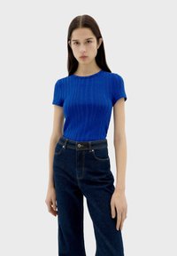 Haut à manches courtes bleu à côtes avec un col rond, associé à un jean en denim foncé taille haute. Le tissu semble lisse avec un léger éclat.