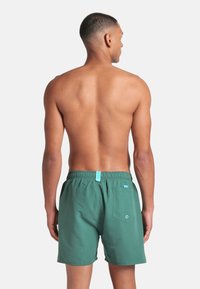 Uomo con capelli corti in piedi di spalle, indossando pantaloni da bagno verdi con una tasca posteriore e un'etichetta blu su una cintura elastica.