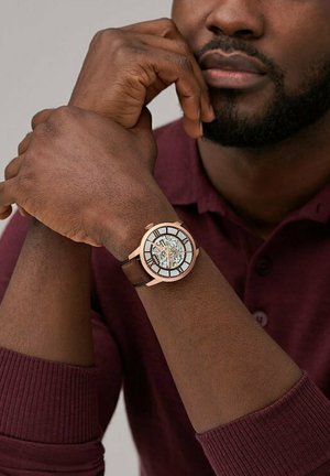 Homme à la peau foncée portant une chemise bordeaux à manches longues montre une montre à bracelet en cuir marron avec un mécanisme d'engrenage visible au poignet.