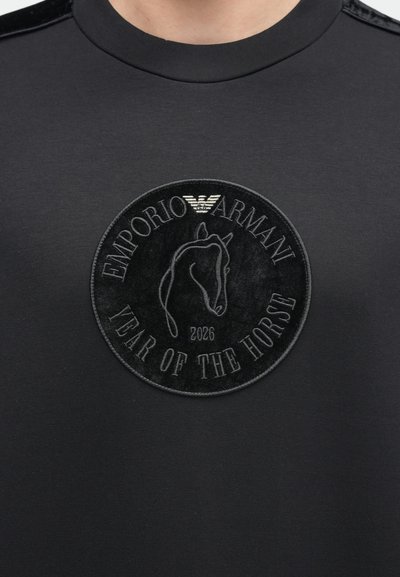Melna kokvilnas T-krekls ar apaļu izceltu izšuvumu ar zirga dizainu un tekstu "EMPORIO ARMANI YEAR OF THE HORSE 2026". Gluda tekstūra.