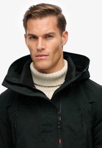 Dunkelgrüne Kapuzenjacke mit Reißverschluss, ausgestattet mit zwei Reißverschlusstaschen und dem Schriftzug "SUPER." Über einem cremefarbenen Rollkragenpullover gestylt.
