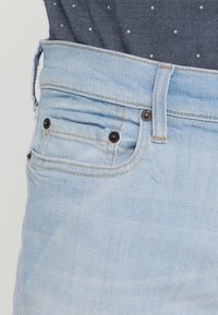 Jeans de mezclilla azul claro con costuras visibles, diseño de cinco bolsillos y cierre de botón. La textura parece suave con un ligero desvanecimiento.