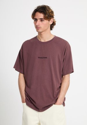Jeune homme aux cheveux ondulés portant un t-shirt ample mauve Volcom et un pantalon clair, les mains dans les poches, regardant légèrement vers la droite.