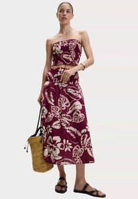 Top corto bustier floreale e gonna lunga abbinata color bordeaux con motivo a foglie bianche; abbinati a una borsa intrecciata in paglia e sandali neri.