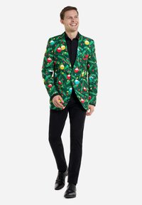 Roheline bleiser, millel on värvilised ornamentide mustrid, punaste, kollaste ja siniste detailidega tekstureeritud taustal, kombineeritud mustade pükste ja kingadega.