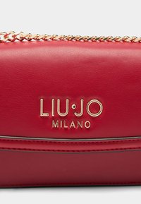 Červená syntetická kožená kabelka se zlatými kovovými doplňky a řetízkovým popruhem, vybavená embosovaným logem a hladkým povrchem.