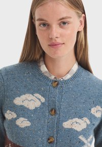 Modrý pletený cardigan se vzorem mraků, kulatým výstřihem a třemi hnědými knoflíky. Texturovaná látka s žíhanými detaily po celém povrchu.