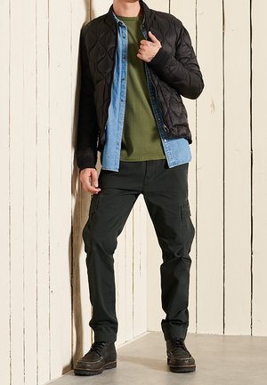 Veste bomber matelassée noire, T-shirt vert, chemise en denim superposée, pantalon cargo foncé et bottes noires, debout contre un mur en bois clair.