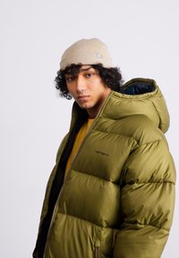 Olive green puffer jacket with a zip front, featuring a hood and a Carhartt logo. Model wears a beige beanie and a yellow shirt underneath.