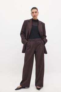 Braun maßgeschneiderte Blazer und weite Hosen aus einem glatten Stoff, kombiniert mit einem schwarzen Rundhalsshirt und spitzen schwarzen Ballerinas.
