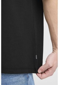 Sort bomulds T-shirt med lige kant, sideslitz og et lille stribet stofmærke nederst. En hånd der holder kanten er synlig.