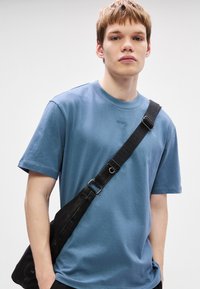 Μπλε βαμβακερό t-shirt με κοντά μανίκια, στρογγυλή λαιμοκοπή και μικρή λεπτομέρεια λογότυπου. Μαύρη τσάντα ώμου με ρυθμιζόμενο λουρί και πόρπη.