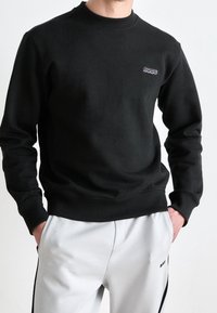 Muž v černém svetru s kulatým výstřihem, na kterém je logo "BOSS" na hrudi, a světle šedých teplákách, s rukama v kapsách.