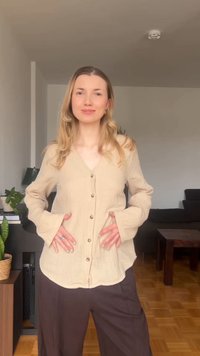 Νεαρή γυναίκα με μακριά ξανθά μαλλιά, ντυμένη με μπεζ πουκάμισο με κουμπιά και σκούρο παντελόνι, στέκεται μέσα σε κλειστό χώρο με τα χέρια στη μέση.