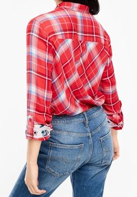 Chemise à carreaux rouge et bleu avec un col à boutons, manches retroussées ornées de accents floraux, associée à un jean en denim bleu taille haute.