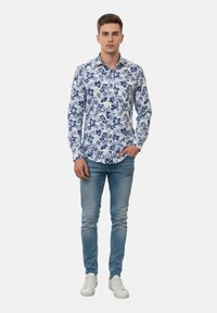 Joven de pie con la mano izquierda en el bolsillo, vistiendo una camisa blanca con patrón floral azul, jeans azul claro y zapatillas blancas.