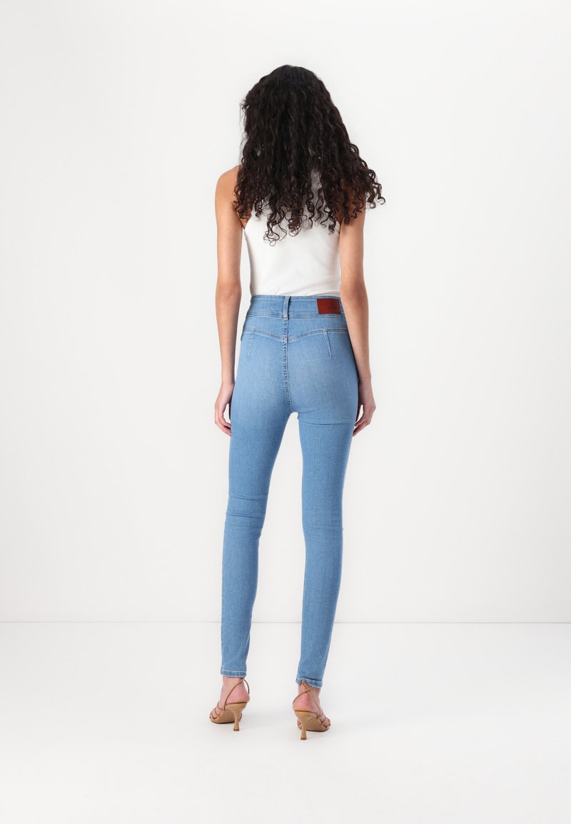 Hoch geschnittene, hellblaue Skinny-Jeans mit einer glatten Textur, ausgestattet mit einem braunen Leder-Patch hinten und Reißverschlüssen an den Knöcheln.