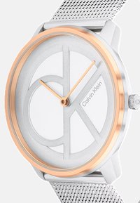 Sidabrinis nerūdijančio plieno laikrodis su tinkleliu, baltas ciferblatas su dideliu išpjovos "CK" dizainu ir rožinės auksinės tonas bezelis bei rodyklės.