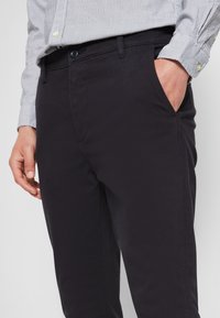 Schwarze Baumwollchinos mit einem eleganten Design, ausgestattet mit einem Knopfverschluss und seitlichen Taschen. Glatte Textur und maßgeschneiderte Passform sind erkennbar.