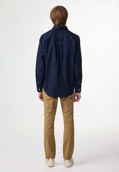 Camicia a maniche lunghe blu navy con colletto a punta, polsini abbottonati e spacchi laterali, abbinata a pantaloni beige chiari e scarpe casual.