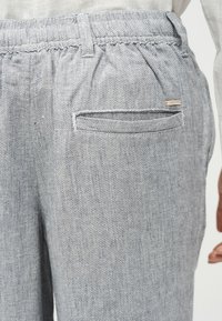 Κοντινό πλάνο σε παντελόνι από ανοιχτό γκρι υφαντό ύφασμα με ελαστική μέση, μια τσέπη με φιτίλι στην πλάτη και μικρή μεταλλική ετικέτα με το λογότυπο.