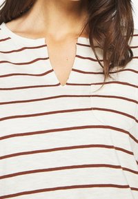 Haut à rayures en v avec un tissu léger. Fond crème avec de fines rayures horizontales marron. Comprend une petite poche poitrine.