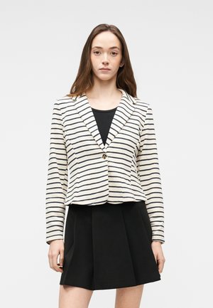 Femme aux longs cheveux bruns portant un blazer blanc à rayures horizontales noires et une jupe noire, debout devant un fond blanc.