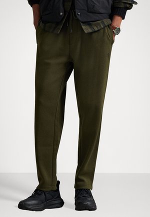 Pantaloni de trening de culoare verde măsliniu, cu picioare conice, talie elastică și buzunare laterale. Asortați cu pantofi sport negri. Textură de material neted.