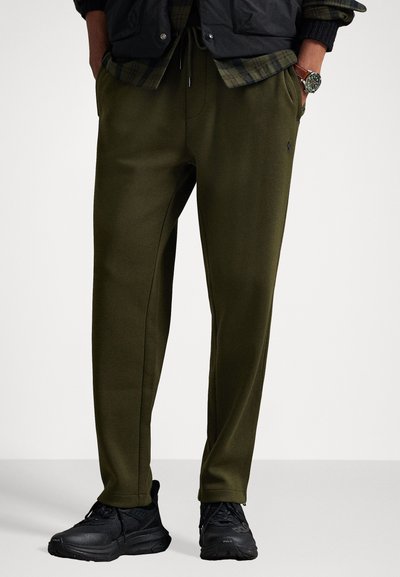 Pantalones de chándal verde oliva con piernas con diseño cónico, cintura elástica y bolsillos laterales. Combinados con zapatos deportivos negros. Textura de tela suave.
