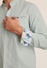 Lichtgroene overhemden met kraag en een borstzak; de manchetten hebben een blauw-wit abstract patroon en limoen groene afwerking. Drukknopen zichtbaar.