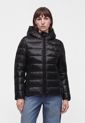 Frau trägt eine schwarze, wattierte Kapuzenjacke mit Frontreißverschluss und blaue Jeans, steht vor einem schlichten hellen Hintergrund.