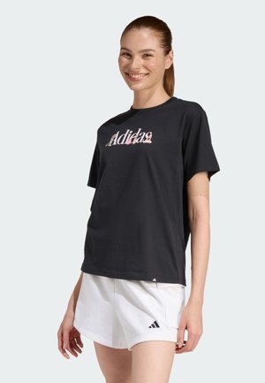 Donna sorridente con capelli rossi, indossa una maglietta nera Adidas e pantaloncini bianchi Adidas, in piedi davanti a uno sfondo chiaro e semplice.