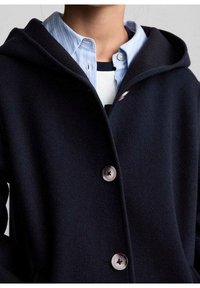 Námořnický kabát s kapucí vyrobený z měkké tkaniny, který má velké knoflíky a volný střih, přehozený přes světle modrou pruhovanou košili s límečkem.