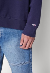 Marineblå genser med ribbestrikkede mansjetter, med en liten flerfarget logo på ermet. Båret med lyseblå denimjeans som viser detaljert tekstur.
